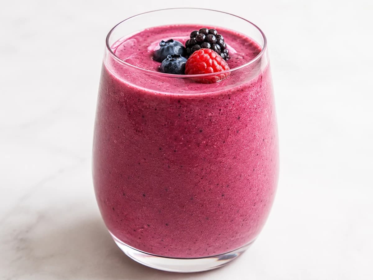 Tall glass of berry smoothie with a few whole berries and a banana beside it; neutral background.