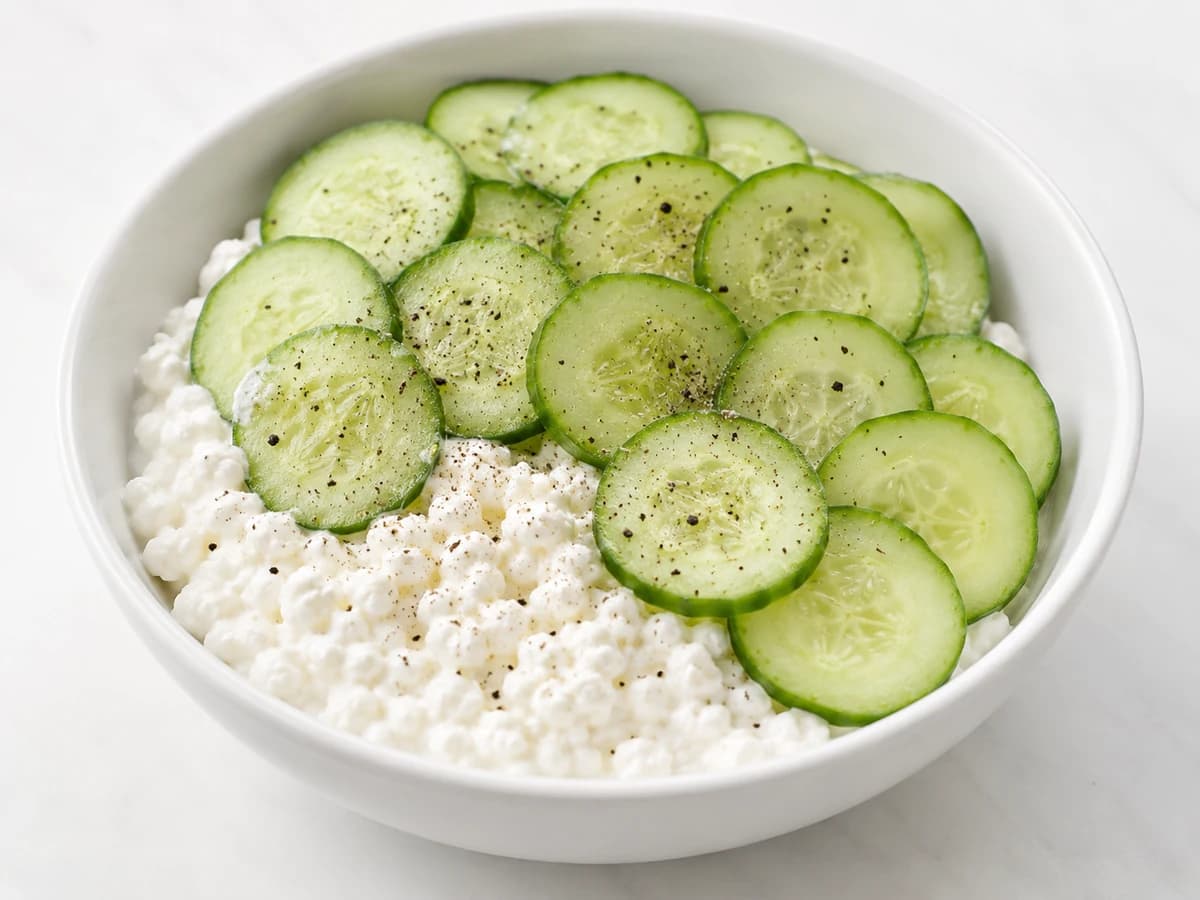Small bowl of cottage cheese with raw cucumber slices and cracked black pepper; top-down.